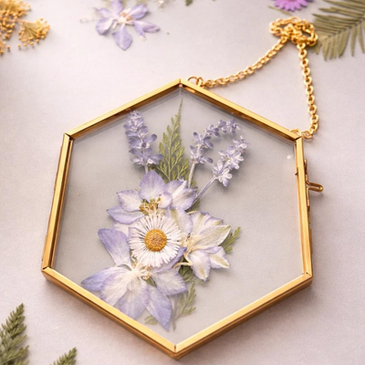 A gold hexagonal glass frame pendant pressed with white daisies, purple delphinium, and greenery, hanging from a delicate gold chain